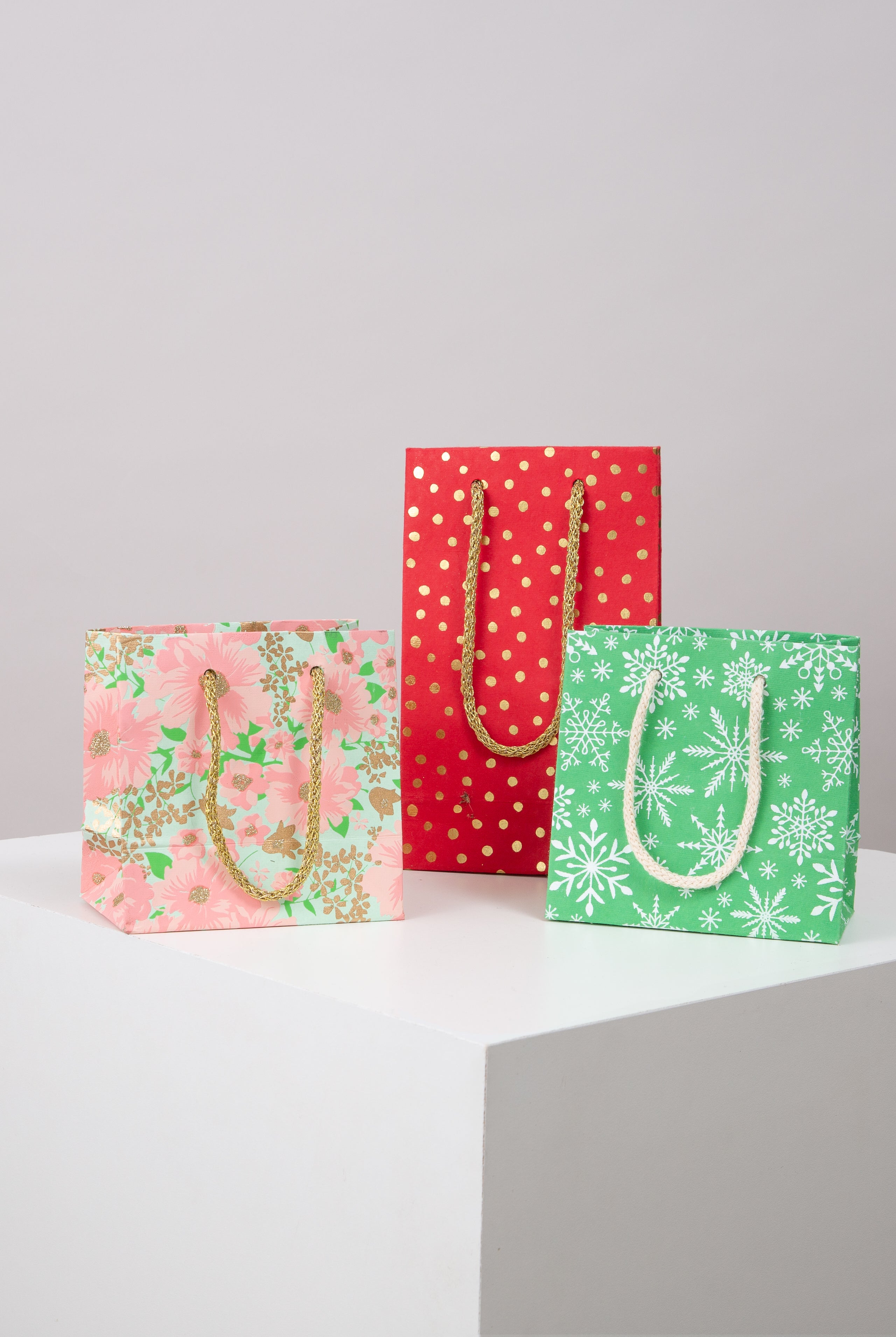 Three examples of the Upcycled Gift Bags are pictured. Two are square, and one is tall and skinny. Each has two woven cord handles in a complementary color attached to the top of the bag. The bags are each a different color and pattern. One has a floral pattern, one is polka dotted, and one has a snowflake pattern.