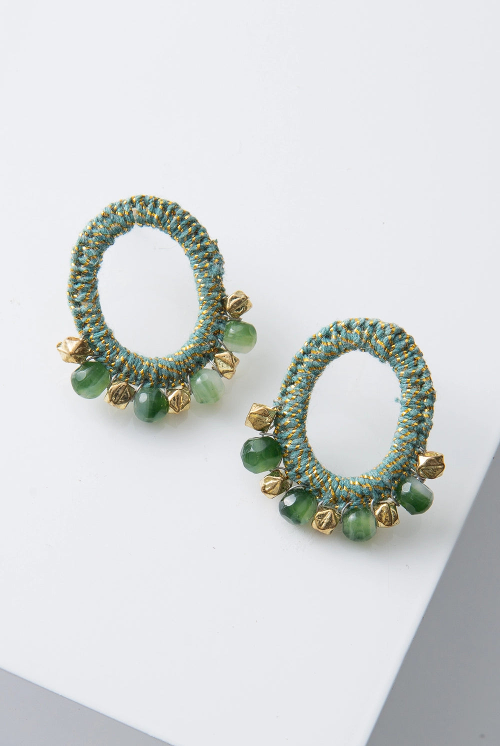 The Anjana Crochet Earrings are mid-size post earrings. The ear post connects to an oval hoop that is wrapped in mint green and metallic gold crocheted thread. Faceted gold metal beads and jade green glass beads are arranged along the bottom of the hoop.