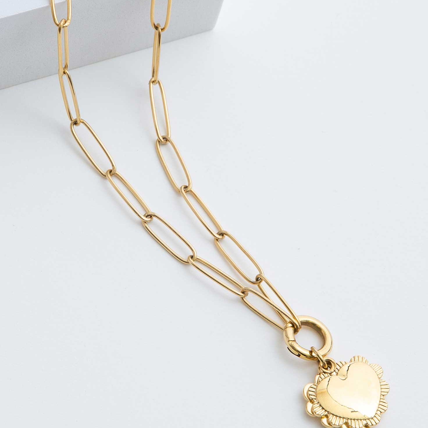 The Ardent Necklace is a gold pendant locket. A heart pendant outlined in a ruffled border hangs from a gold chain. The chain has elongated oval-shaped links.