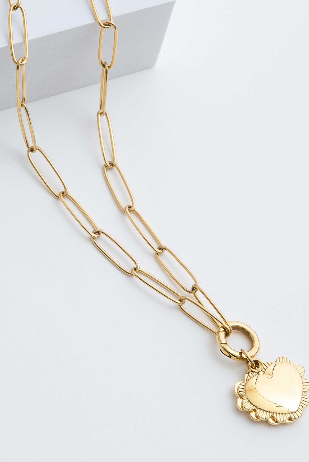 The Ardent Necklace is a gold pendant locket. A heart pendant outlined in a ruffled border hangs from a gold chain. The chain has elongated oval-shaped links.