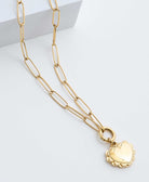 The Ardent Necklace is a gold pendant locket. A heart pendant outlined in a ruffled border hangs from a gold chain. The chain has elongated oval-shaped links.