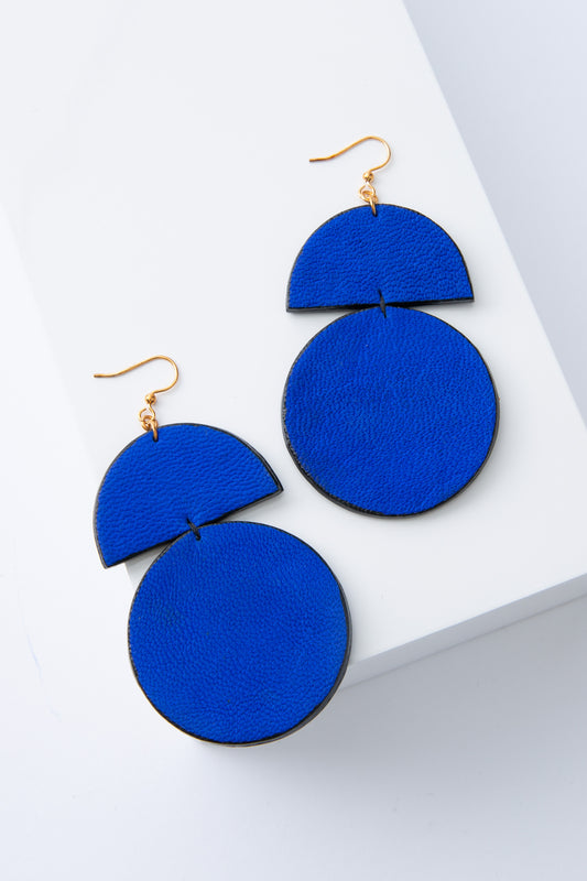 The Assembly Earrings are large statement earrings composed of bright cobalt blue leather. At the top of the earrings is a leather semi-circle. A leather circle is attached below this, creating a geometric and sculptural look.