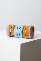 This is a tubular shaped pouch composed of brightly patterned woven cotton. The pouch is wider than it is tall. A zipper stretches along the top of the pouch. The fabric features stripes of mustard yellow, light blue, aqua, pink, and navy, as well as stripes with a traditional ikat pattern.