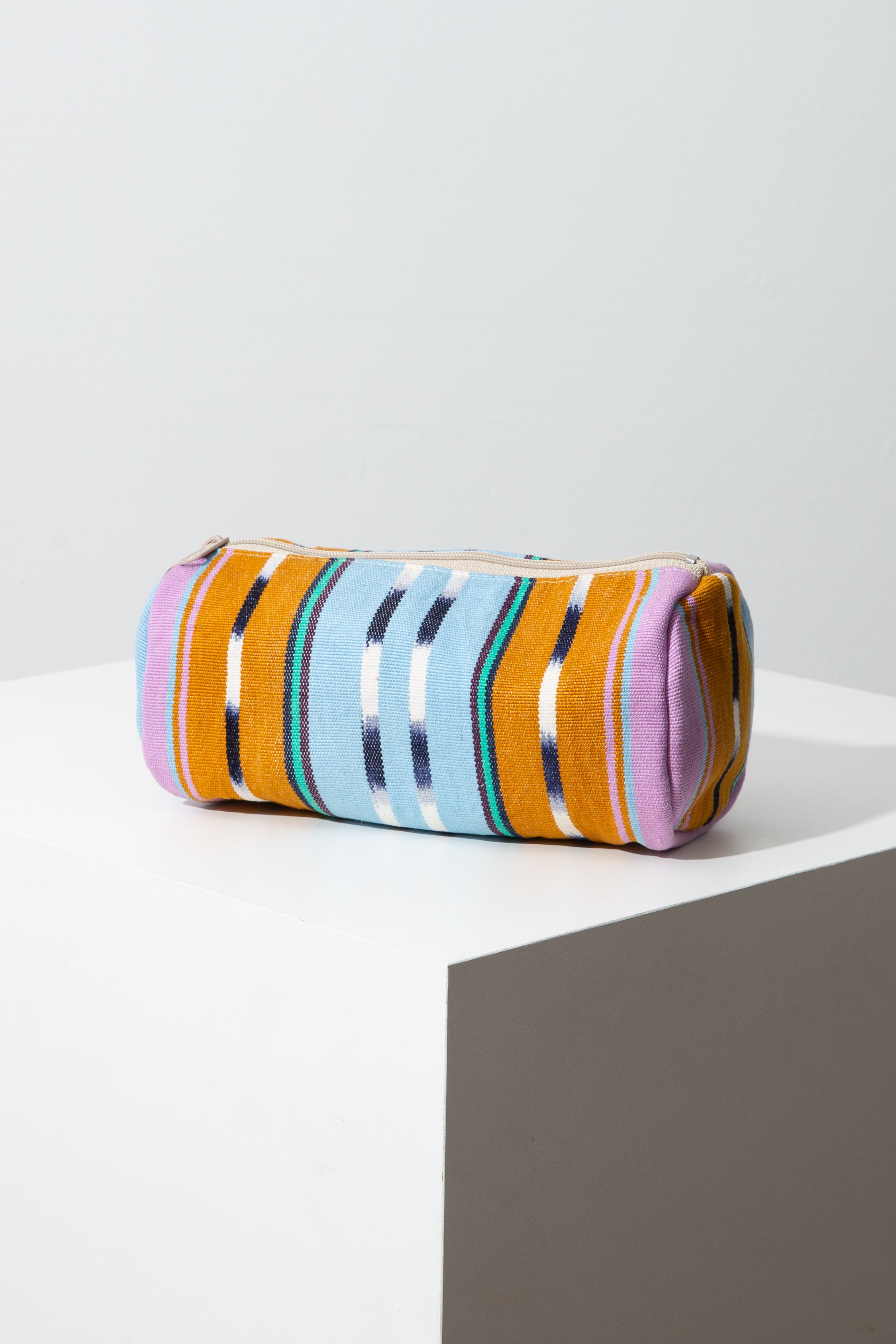 This is a tubular shaped pouch composed of brightly patterned woven cotton. The pouch is wider than it is tall. A zipper stretches along the top of the pouch. The fabric features stripes of mustard yellow, light blue, aqua, pink, and navy, as well as stripes with a traditional ikat pattern.