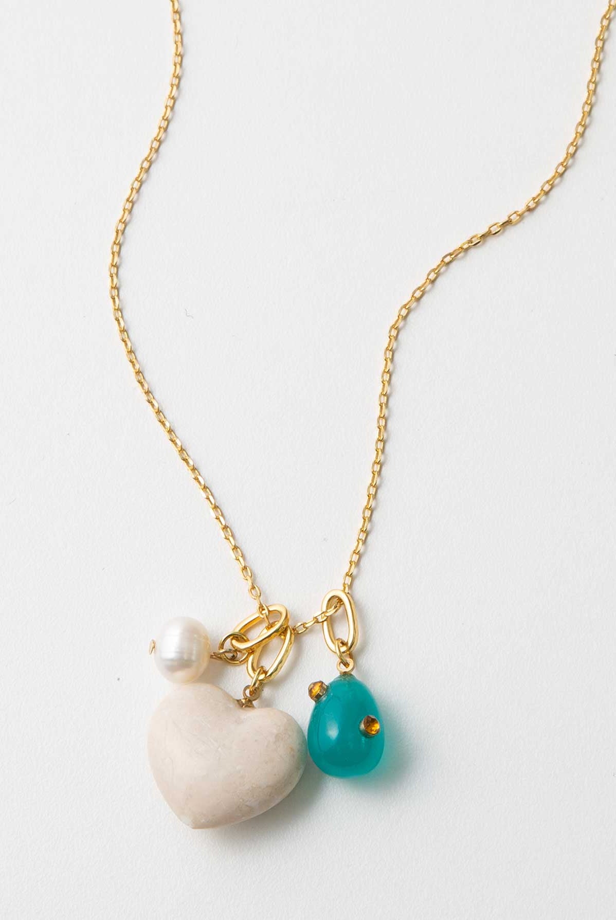 The Big Heart Charm Necklace has a dainty gold chain with three charms. One is  small round pearl, one is a peach colored marble heart, and one is a turquoise glass teardrop. Each hangs from a gold ring.