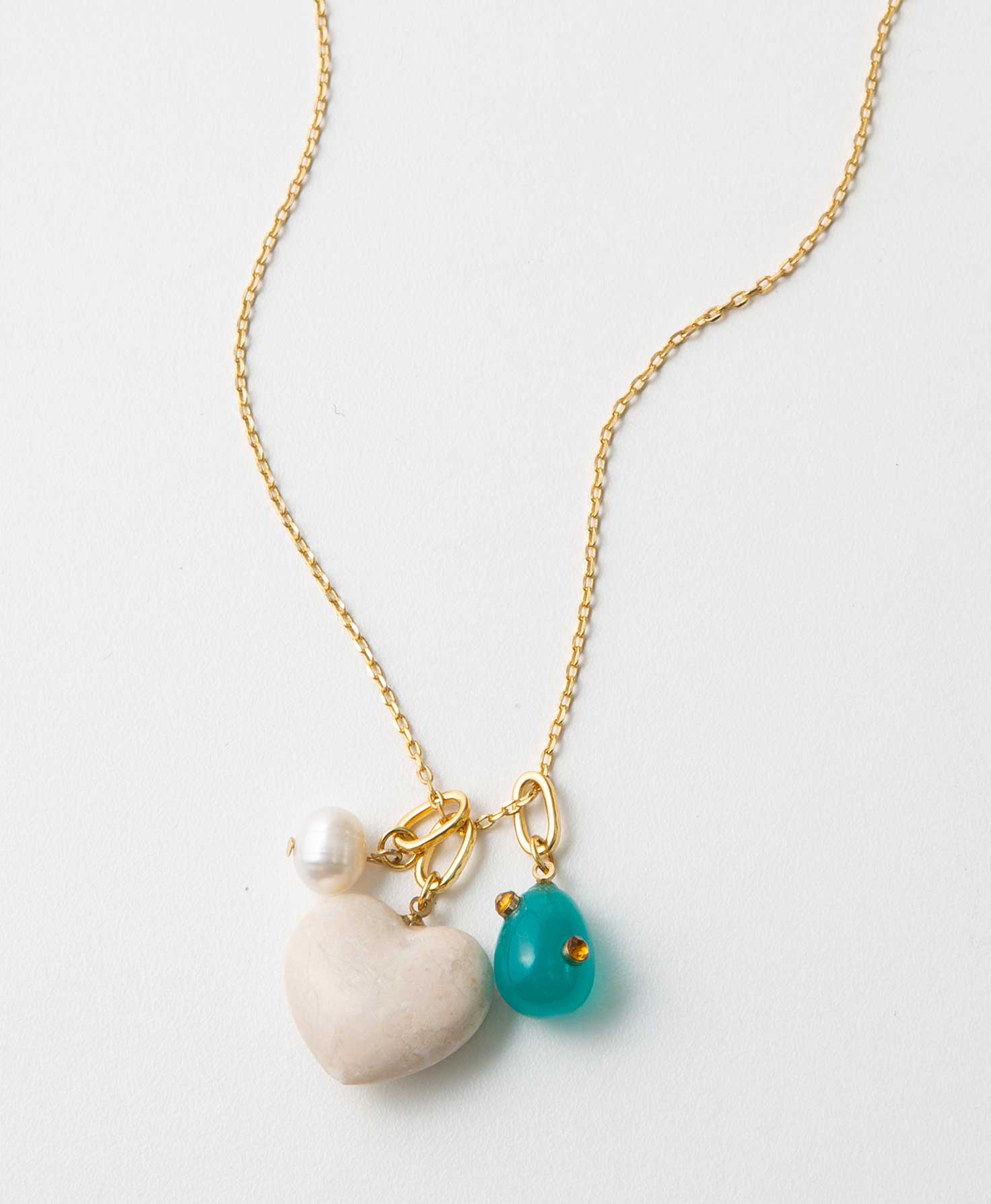 The Big Heart Charm Necklace has a dainty gold chain with three charms. One is  small round pearl, one is a peach colored marble heart, and one is a turquoise glass teardrop. Each hangs from a gold ring.