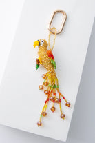 A three-dimensional beaded bird hangs from a brass oval keyring. The bird is primarily composed of translucent yellow beads. It has a beak, eyes, wings, and tail feathers composed of different colored beads. The beak is gold and the wings are red, yellow, and green. A long beaded fringe hangs down, forming the tail feathers.