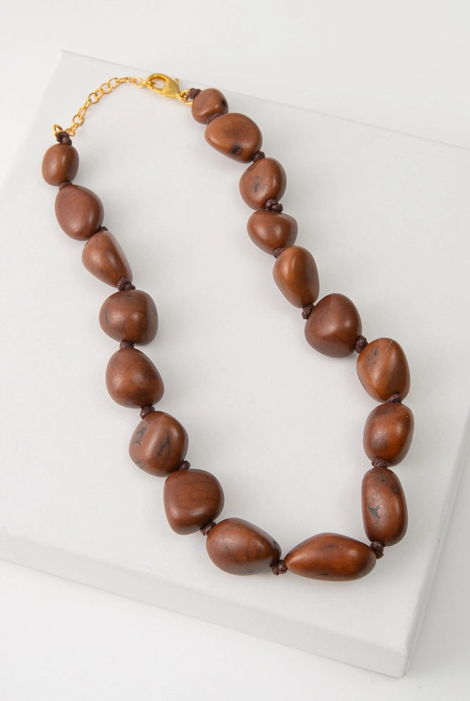 The Bisola Bauble Necklace features a single strand of shiny brown seeds called Bisola Seeds. The seeds each have their own unique shapes and natural striations. The seeds closest to the necklace clasps are the smallest, and they gradually increase in size towards the bottom of the necklace. The necklace is finished with a gold chain clasp.