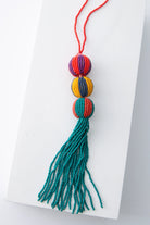 This bag charm has a long strap composed of a strand of red beads. This strap can be used to attach the charm to various items, including a bag strap. The charm itself is composed of three beaded spheres stacked on top of one another, with a long, turquoise beaded fringe at the bottom. Each sphere features two colors of beads that alternate to form a stripe pattern similar to a beach ball. The top sphere is purple and red. The middle sphere is yellow and navy. And the bottom sphere is turquoise and orange.