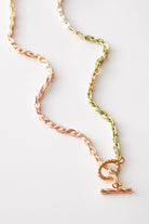 The Braided Gradient Necklace is composed of a gold link chain interwoven with colored thread. The thread changes color from pink to green as it moves along the necklace. There is a chunky gold toggle clasp at the front of the necklace.