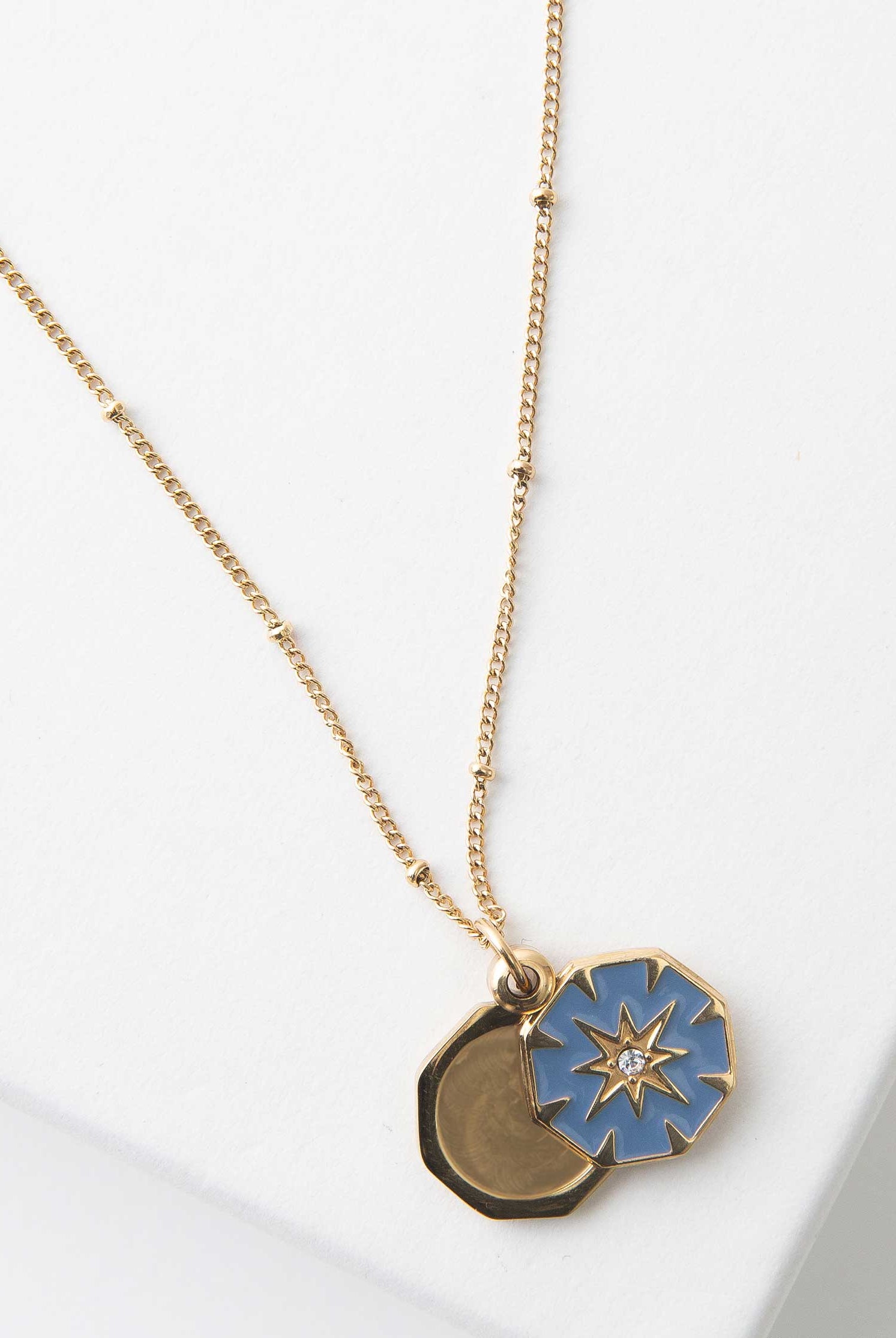 The Bright Star Locket has a gold chain accented with tiny gold beads. The locket is a gold, octogon-shaped pendant. The front panel is filed with blue enamel, and there is a gold starburst design in the middle accented with a clear stone. The front panel swings open to reveal the pendant's interior, where a photograph can be placed.
