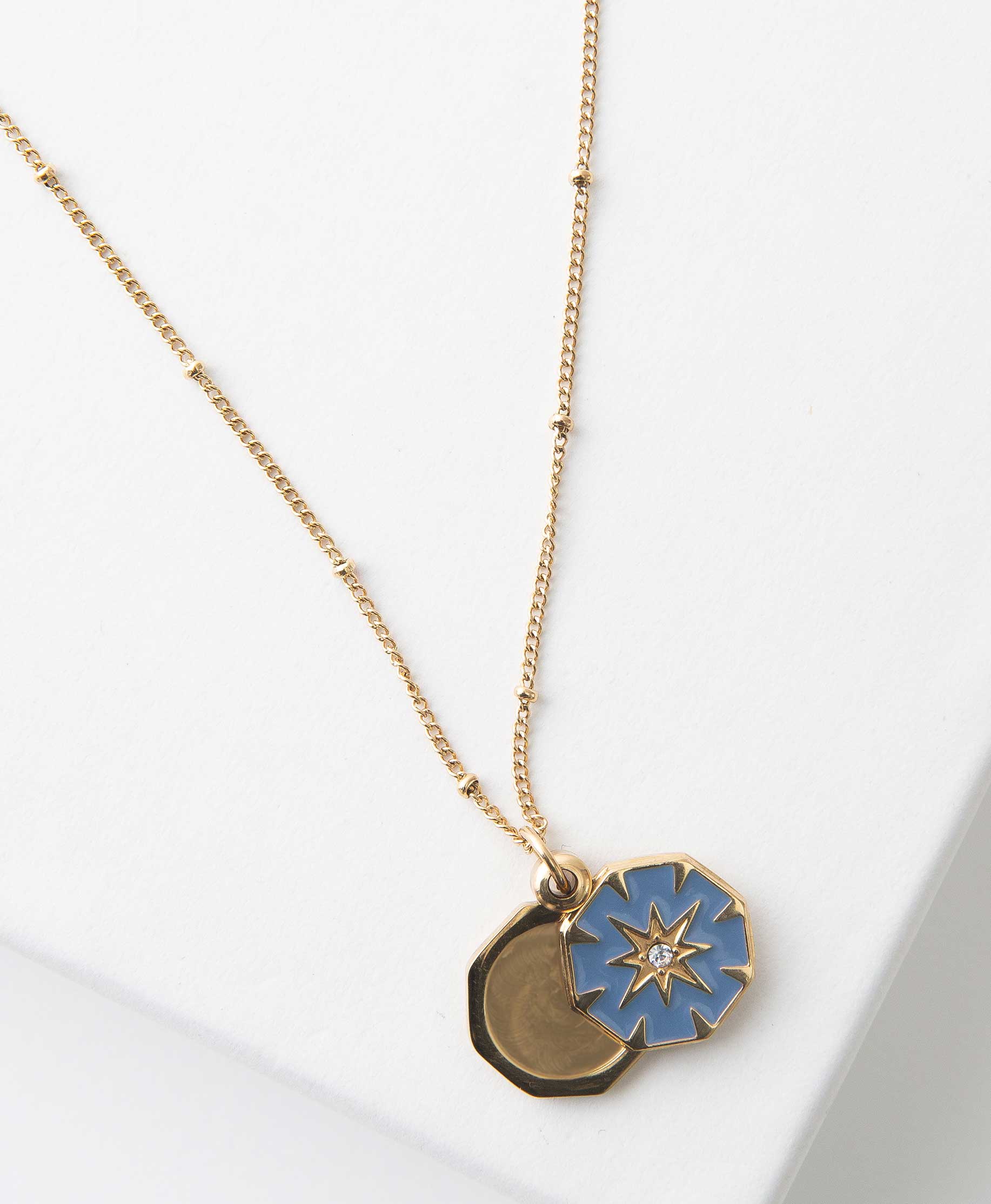 The Bright Star Locket has a gold chain accented with tiny gold beads. The locket is a gold, octogon-shaped pendant. The front panel is filed with blue enamel, and there is a gold starburst design in the middle accented with a clear stone. The front panel swings open to reveal the pendant's interior, where a photograph can be placed.