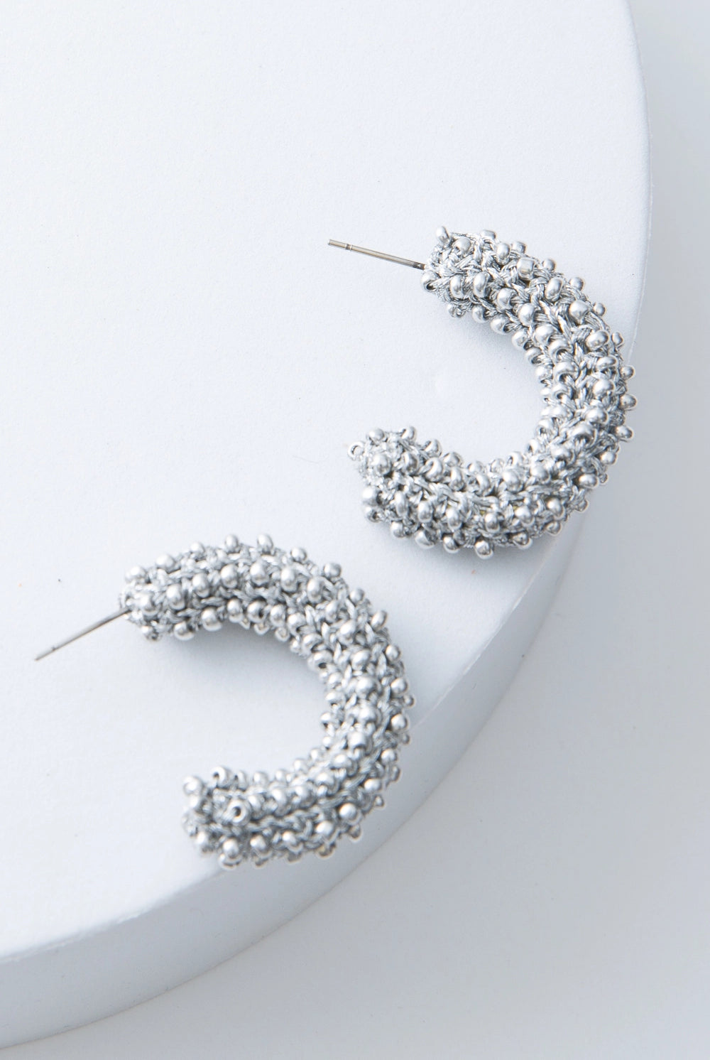 These are silver demi-hoop earrings that are open at the back. They are composed of silver thread and silver beads crocheted together to form a chunky hoop.