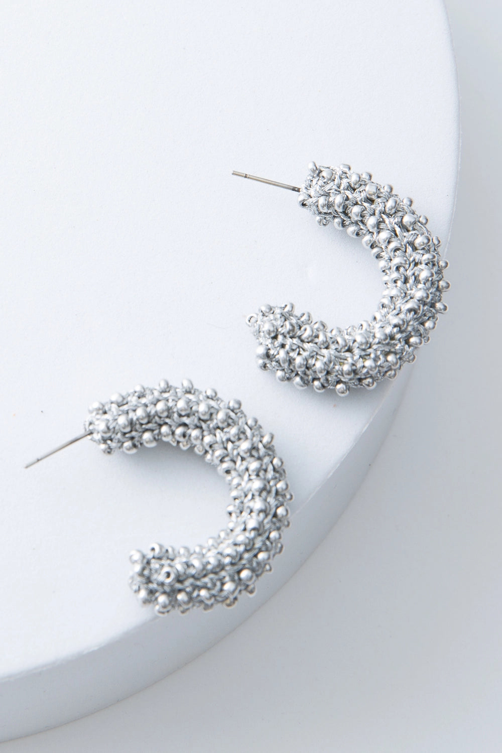 These are silver demi-hoop earrings that are open at the back. They are composed of silver thread and silver beads crocheted together to form a chunky hoop.