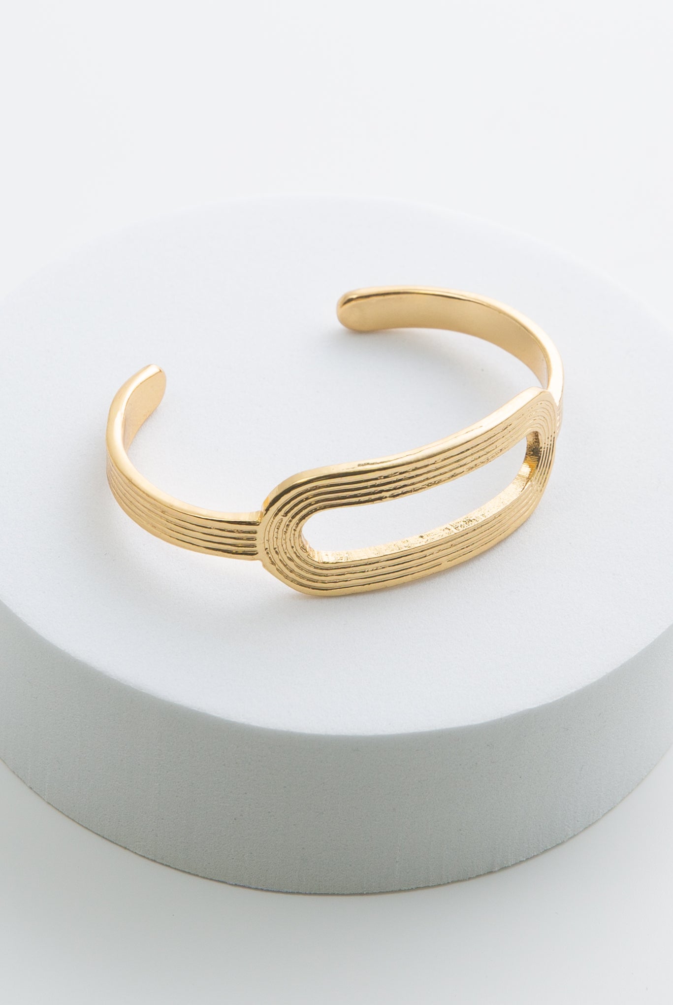 The Circuit Cuff is a thin gold metal bracelet with an opening at the back where the bracelet can be slid onto the wrist. In the center of the bracelet is a horizontal oval made from the same gold metal that is hollow in the middle. There are horizontal lines etched all along the metal, giving the bracelet a ribbed look.