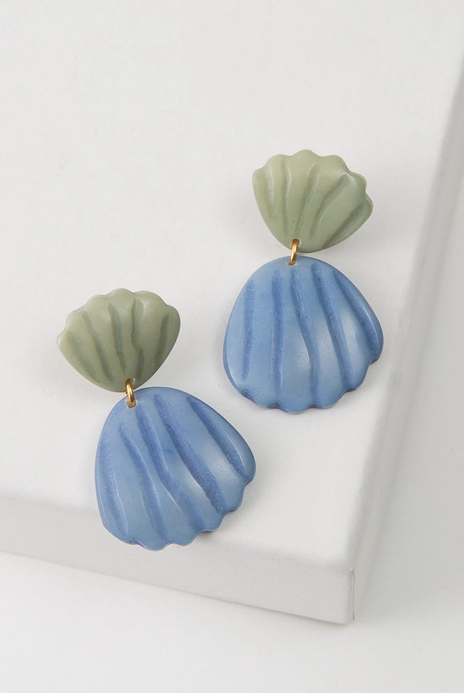 The Coastal Carved Earrings are composed of carved tagua seed. A small shell-shaped tagua piece dyed sage green is attached to an ear post. A larger shell-shaped tagua piece dyed blue hangs down below, connected to the smaller shell with a gold jump ring.