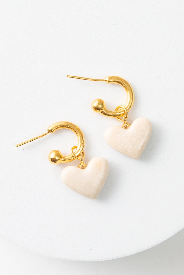 The Convertible Heart Huggies are small gold hoop earrings. There is a cream marble heart charm dangling from the hoops. Each hoop has a gold ball at the end to hold the charm in place. When the earrings are not being worn, the heart charms can slide off for a different look.