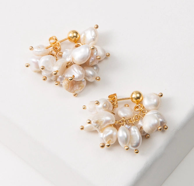 The Convertible Pearl Cluster Earrings are composed of round gold studs with a chain of dangling pearls attached to them. In this image, the earrings are shown with the pearls hooked to the back of the earrings, forming a small hoop. The pearls have a natural look and vary in shape and color.