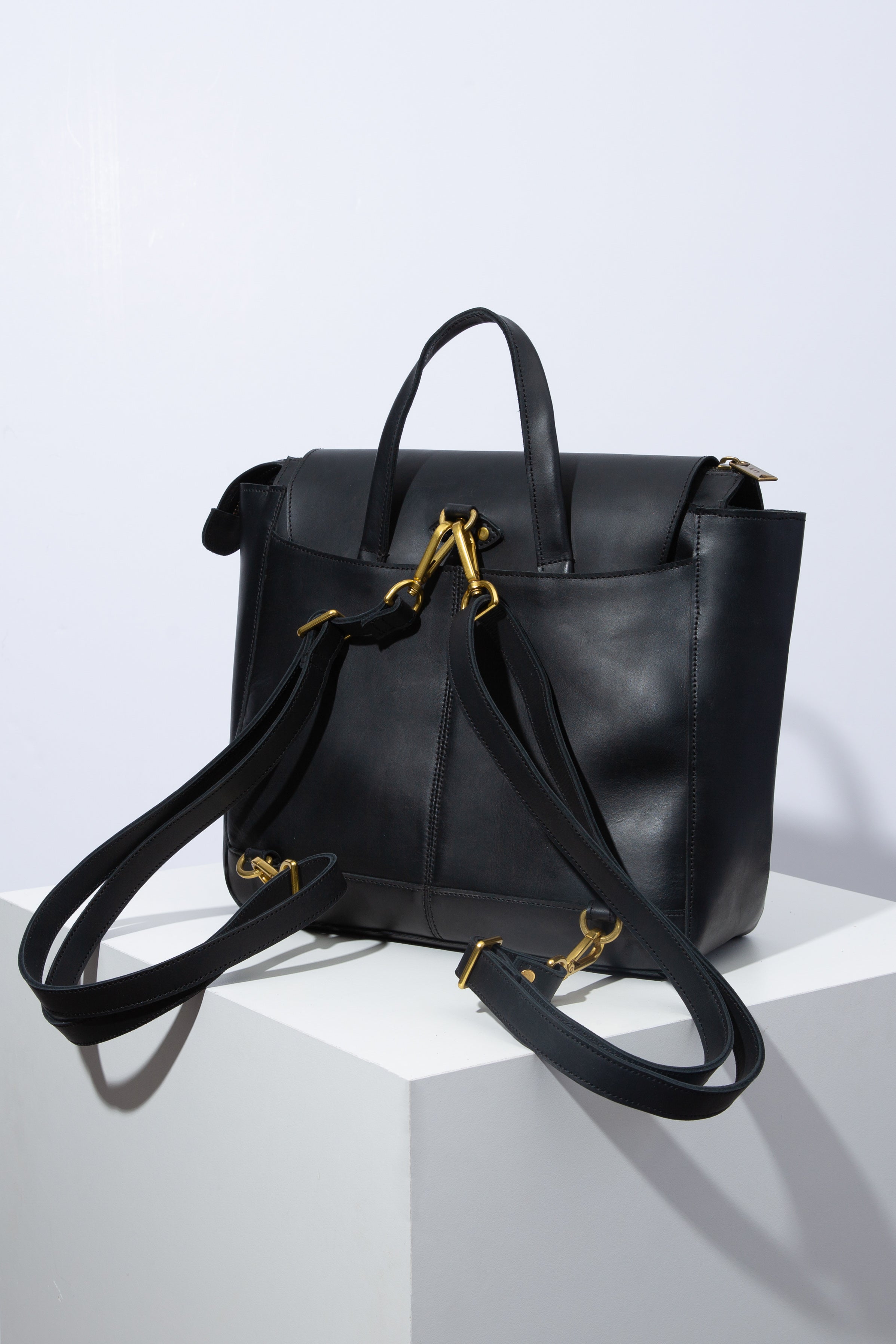 The back of the Debut Backpack is shown. It is styled as a backpack with two leather straps attached to the back of the bag by brass clasps. There is a large slot pocket across the entire back of the bag.