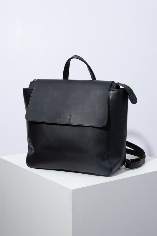 The Debut Backpack is a square leather bag with a wide base that allows it to sit upright. It is composed entirely of glossy black leather. There is a zipper closure across the entire bag. A black leather flap folds over the front of the bag, securing it further. The bag has a short black leather handle at the top, as well as a long, adjustable leather strap that allows the bag to be styled as a shoulder bag or backpack.