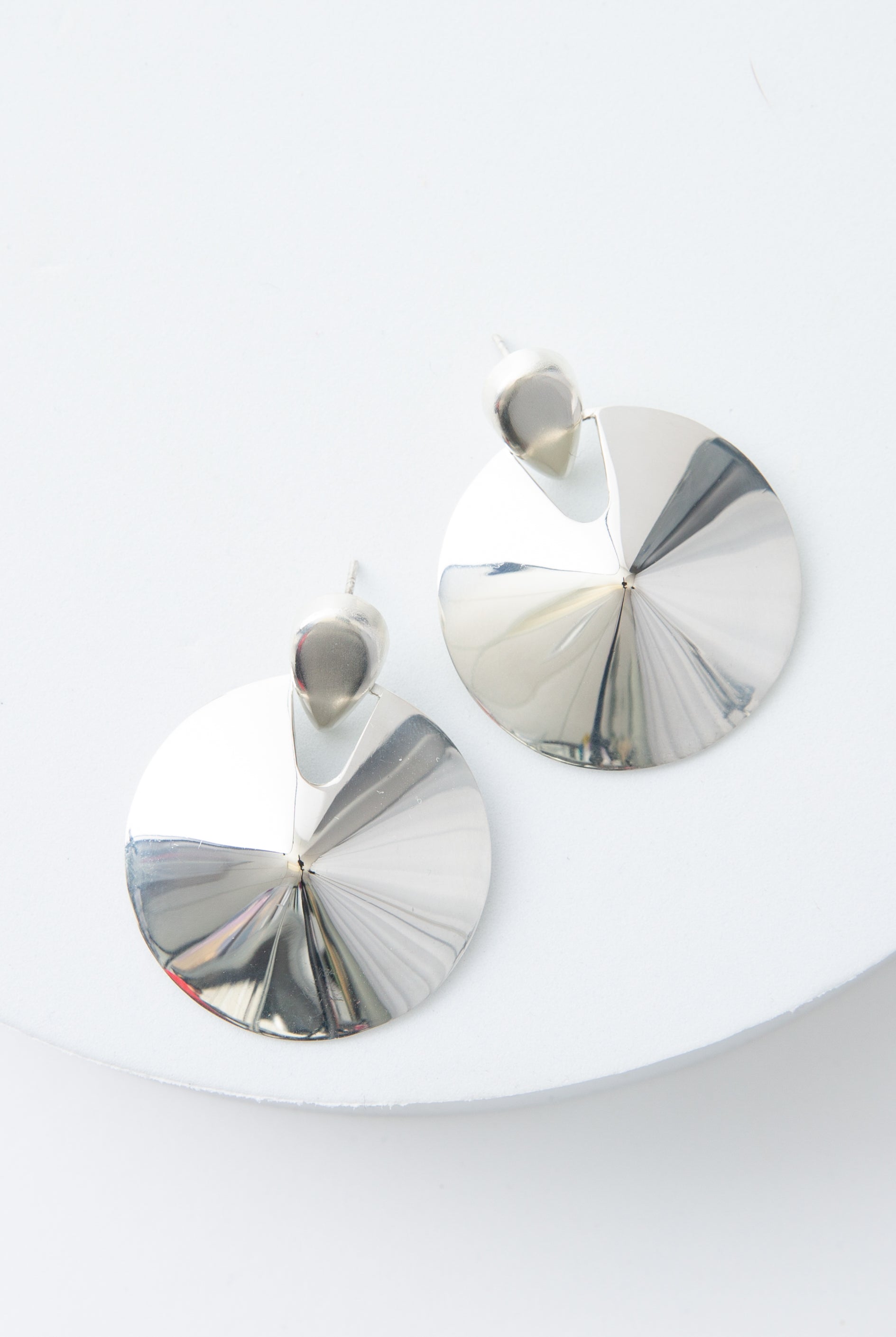 The Defender Earrings are circular disc earrings composed of shining silver. The silver discs are attached to silver ear posts. The discs slope up towards the middle, creating a raised center.