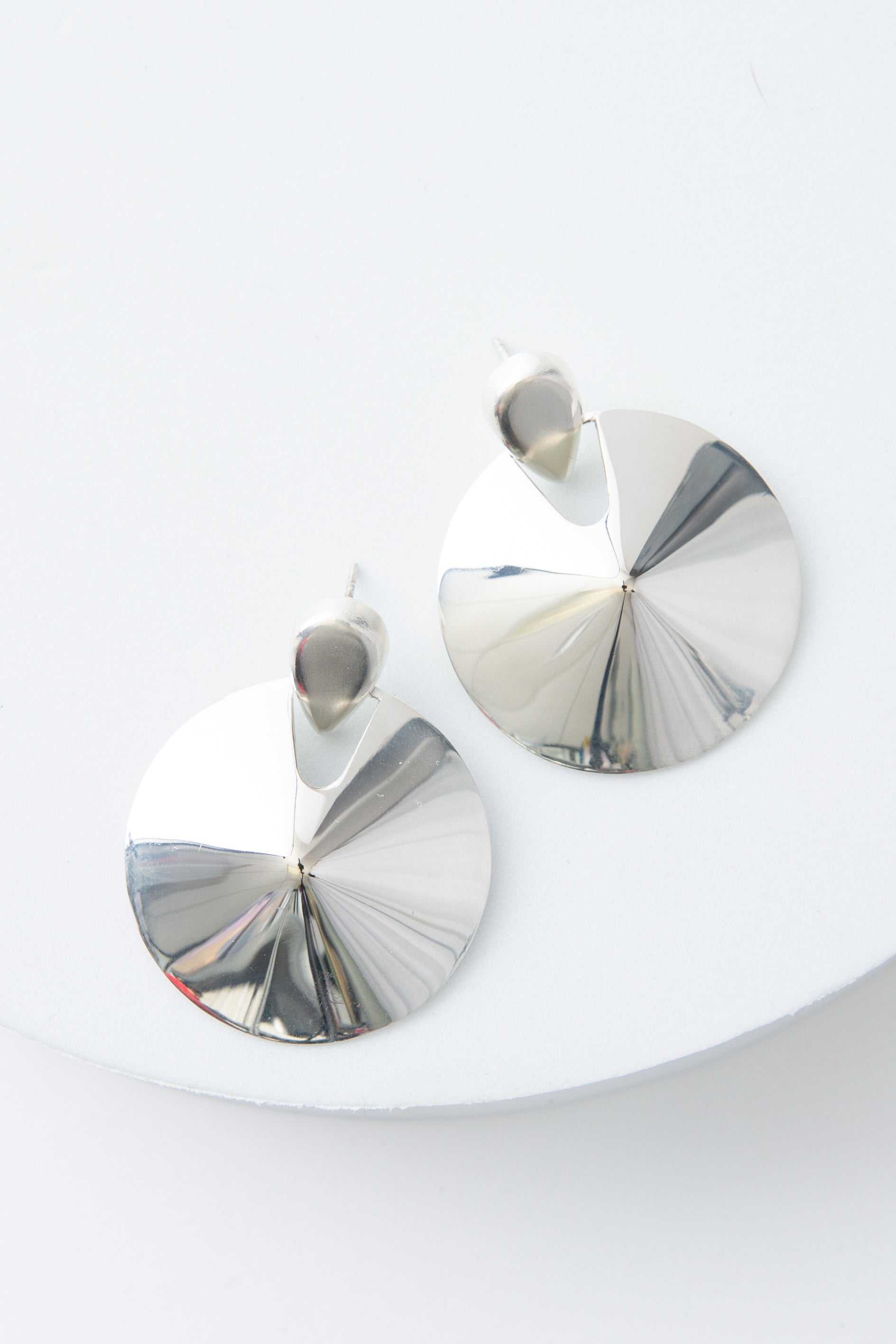 The Defender Earrings are circular disc earrings composed of shining silver. The silver discs are attached to silver ear posts. The discs slope up towards the middle, creating a raised center.