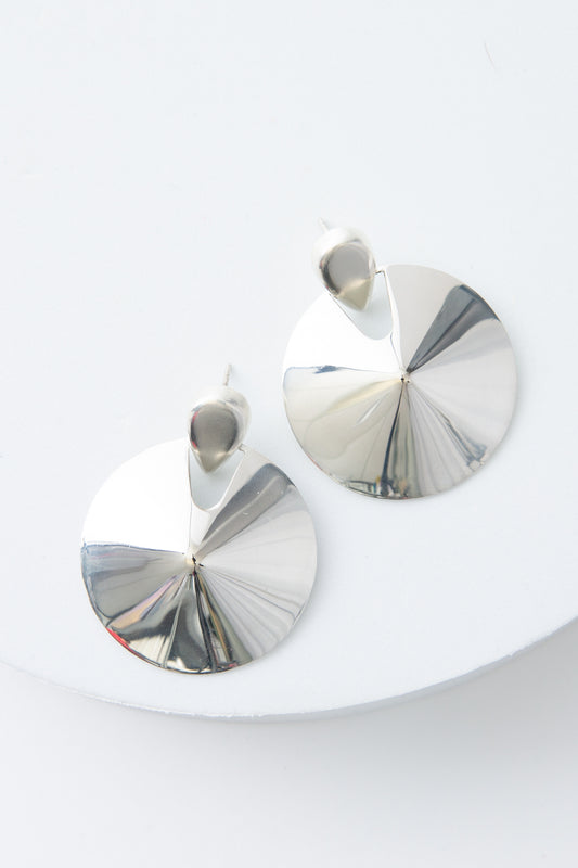 The Defender Earrings are circular disc earrings composed of shining silver. The silver discs are attached to silver ear posts. The discs slope up towards the middle, creating a raised center. 