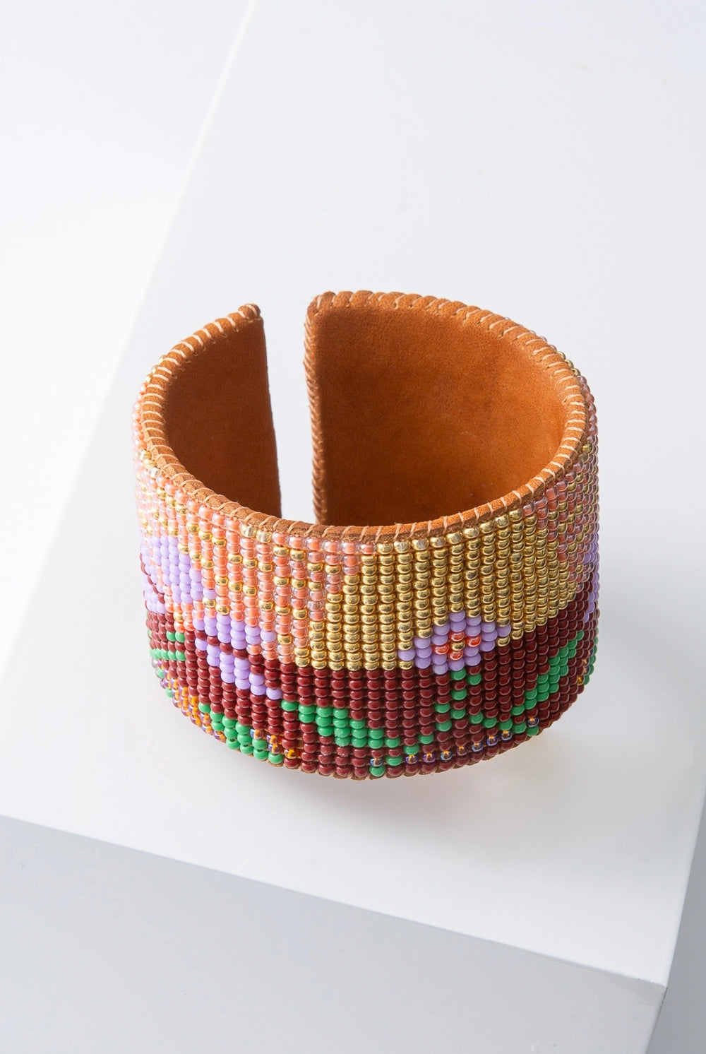 This is a wide, flexible beaded cuff. Soft leather is stitched with hundreds of tiny glass beads to form a desert landscape pattern. The background of the cuff is composed of rust, gold and pink beads that resemble a sunset. Lavender and green beads form flowers and leaves that form the foreground of the landscape.