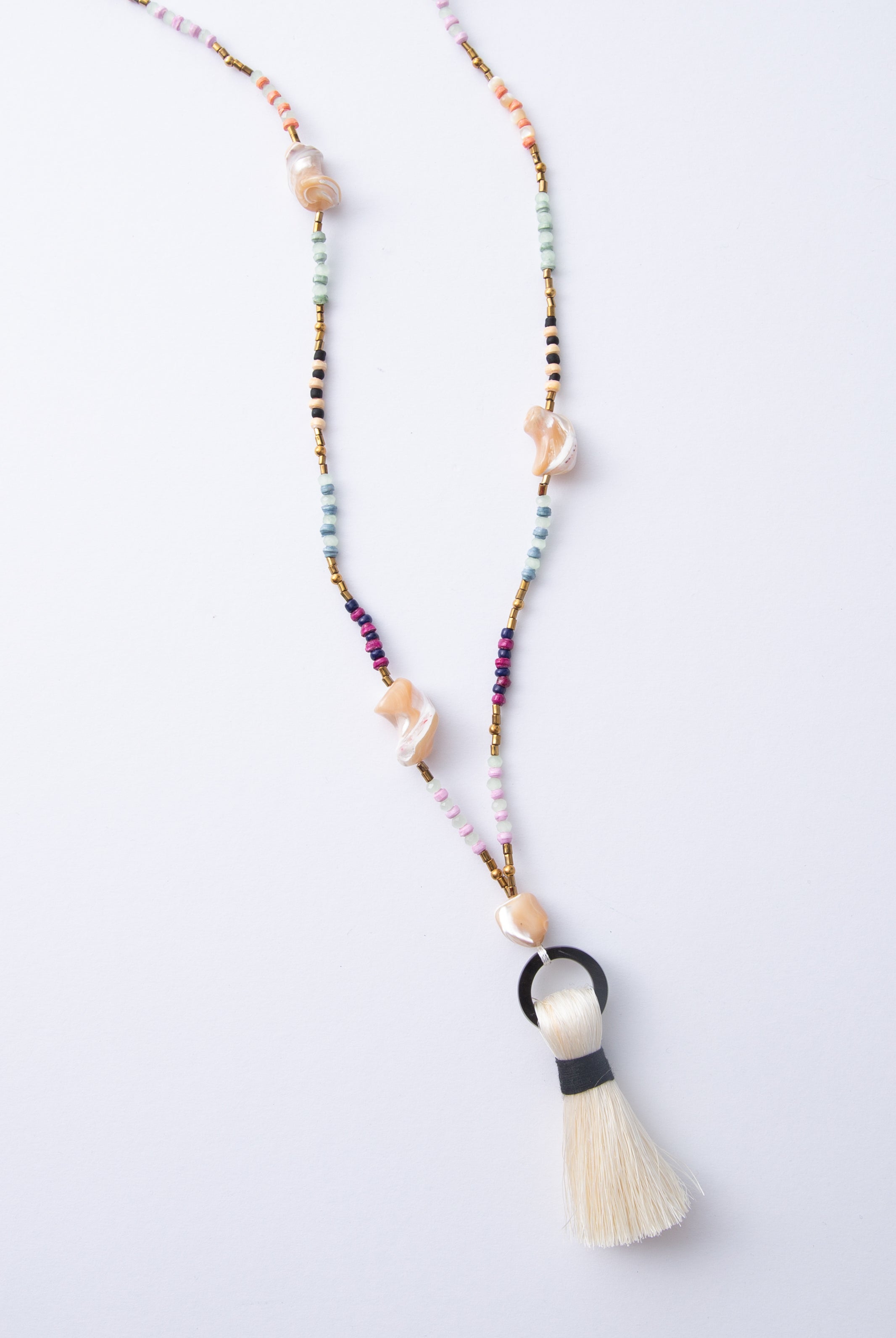 The Dorah Necklace is a lariat-style necklace composed of beads of many different materials and colors. Small paper beads in shades of mint, purple, and cream alternate with small brass beads, glass beads, and large freshwater pearl beads with an irregular, natural shape. At the bottom of the necklace is a horn circle with a cream thread tassel hanging down.