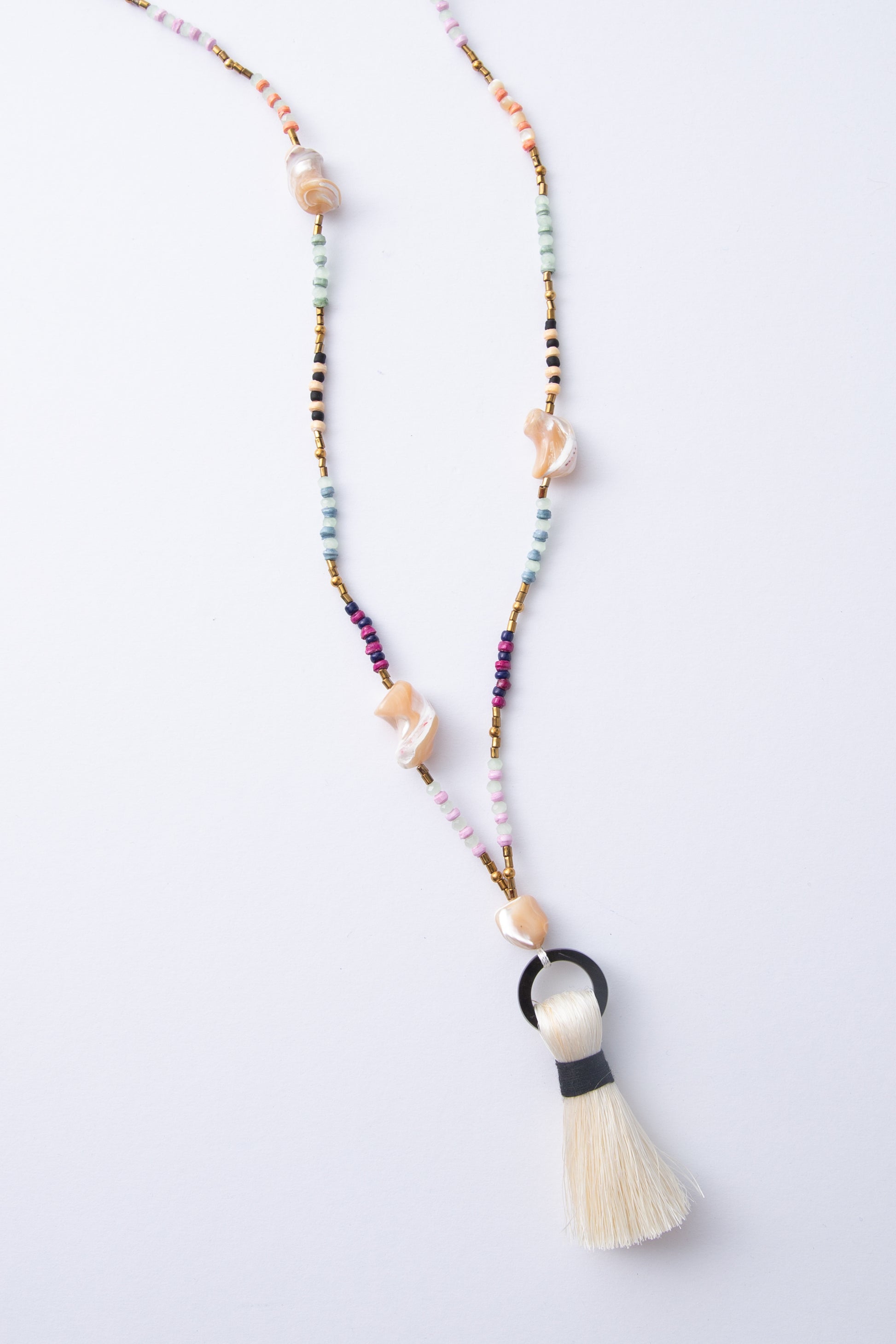The Dorah Necklace is a lariat-style necklace composed of beads of many different materials and colors. Small paper beads in shades of mint, purple, and cream alternate with small brass beads, glass beads, and large freshwater pearl beads with an irregular, natural shape. At the bottom of the necklace is a horn circle with a cream thread tassel hanging down.