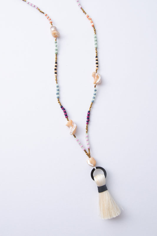 The Dorah Necklace is a lariat-style necklace composed of beads of many different materials and colors. Small paper beads in shades of mint, purple, and cream alternate with small brass beads, glass beads, and large freshwater pearl beads with an irregular, natural shape. At the bottom of the necklace is a horn circle with a cream thread tassel hanging down.