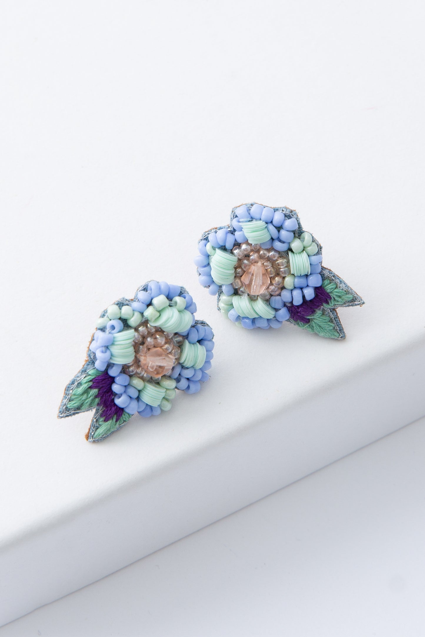 The Forget Me Knot Earrings are post style earrings shaped like blue flowers. The flower is composed of tiny glass beads and sequins in shades of light blue and mint. A cluster of gold beads and a blue colored faceted glass beads form the center of the flower. Two dainty leaves are formed from green and purple thread.