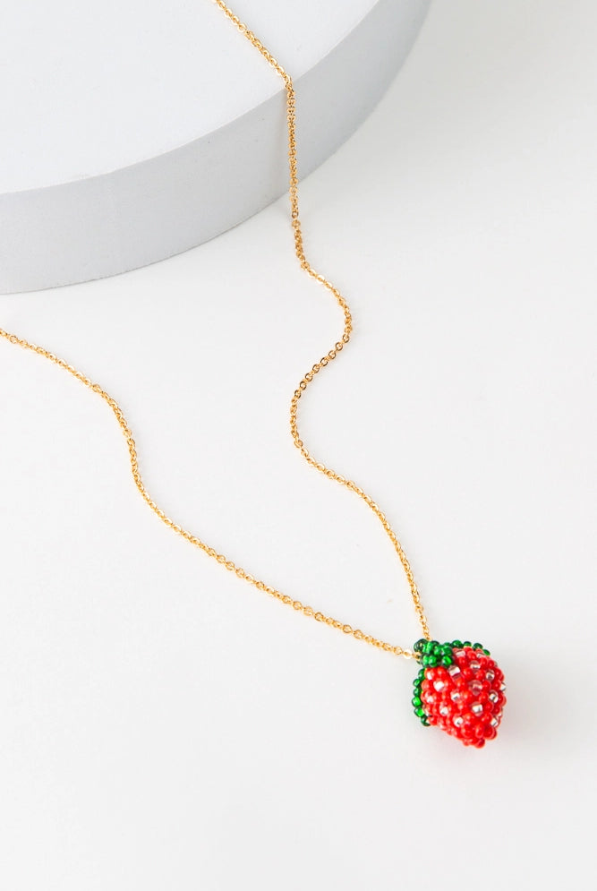 The Frecita Necklace has a dainty gold chain. At the base of the necklace is a tiny strawberry charm composed entirely of translucent glass beads.