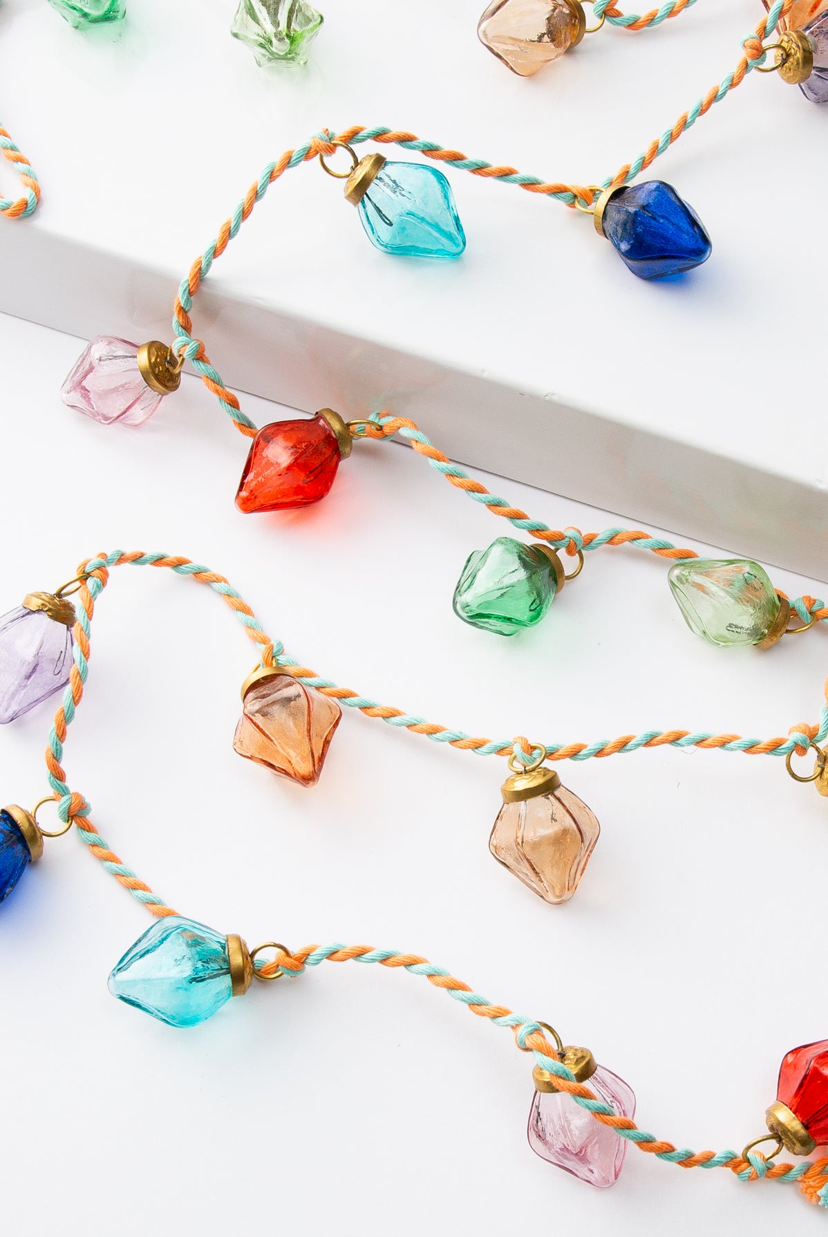 The Glass Bulb Garland lays draped on a white background. The garland is composed of mint and orange cotton thread twisted together. Every few inches there is a large glass bulb knotted onto the thread. The bulbs are a rainbow of colors, including shades of blue, pink, purple, orange, yellow, red, and green. The bulbs have a brass metal top where they are attached to the garland.