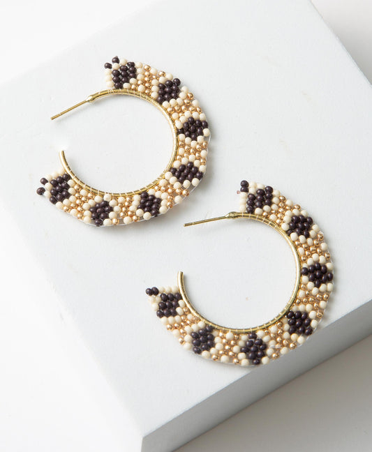 The Glimmer Hoops are composed of a brass metal hoop. Rows of glass beads in shades of gold, white, and black extend out from the hoop. The beads form a geometric diamond pattern. 