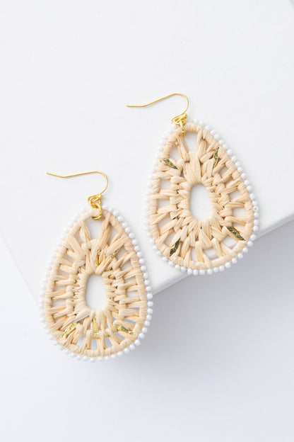 The Goa Earrings are tear-drop shaped earrings composed of woven raffia with an outline of white glass beads. The raffia has a lace-like pattern and is open in the center. The large teardrops hang from brass ear wires.