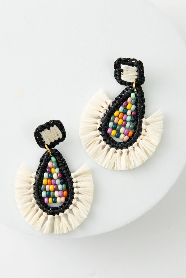 The Gulal Earrings are large teardrop chandelier earrings. The diamond shaped posts are wrapped in black and white raffia. A teardrop shape hangs down below the posts. The exterior rim is wrapped in black raffia, and there is white raffia fringe along the bottom. The interior is composed of rainbow colored glass beads.