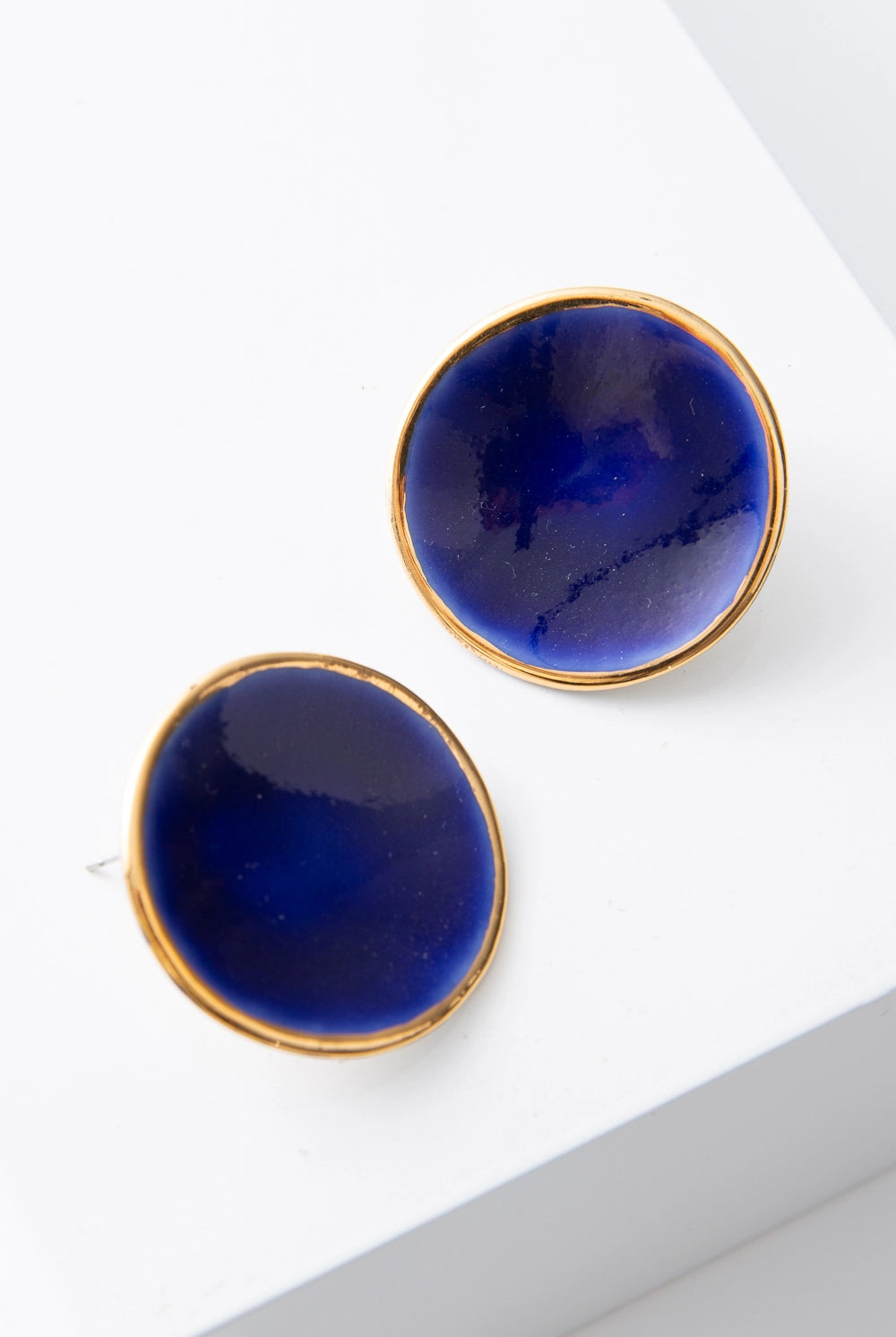 The Halo Earrings are flat, disc-shaped post earrings composed of deep blue porcelain. The discs are outlined in a ring of shining gold. The porcelain has natural variations and subtle markings, giving it a handmade look.