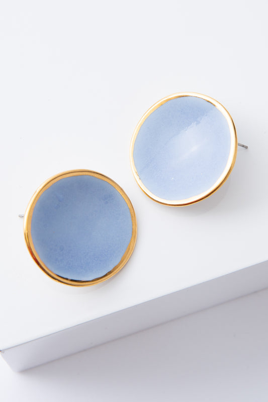 The Halo Earrings are flat, disc-shaped post earrings composed of light blue porcelain. The discs are outlined in a ring of shining gold. The porcelain has natural variations and subtle markings, giving it a handmade look.