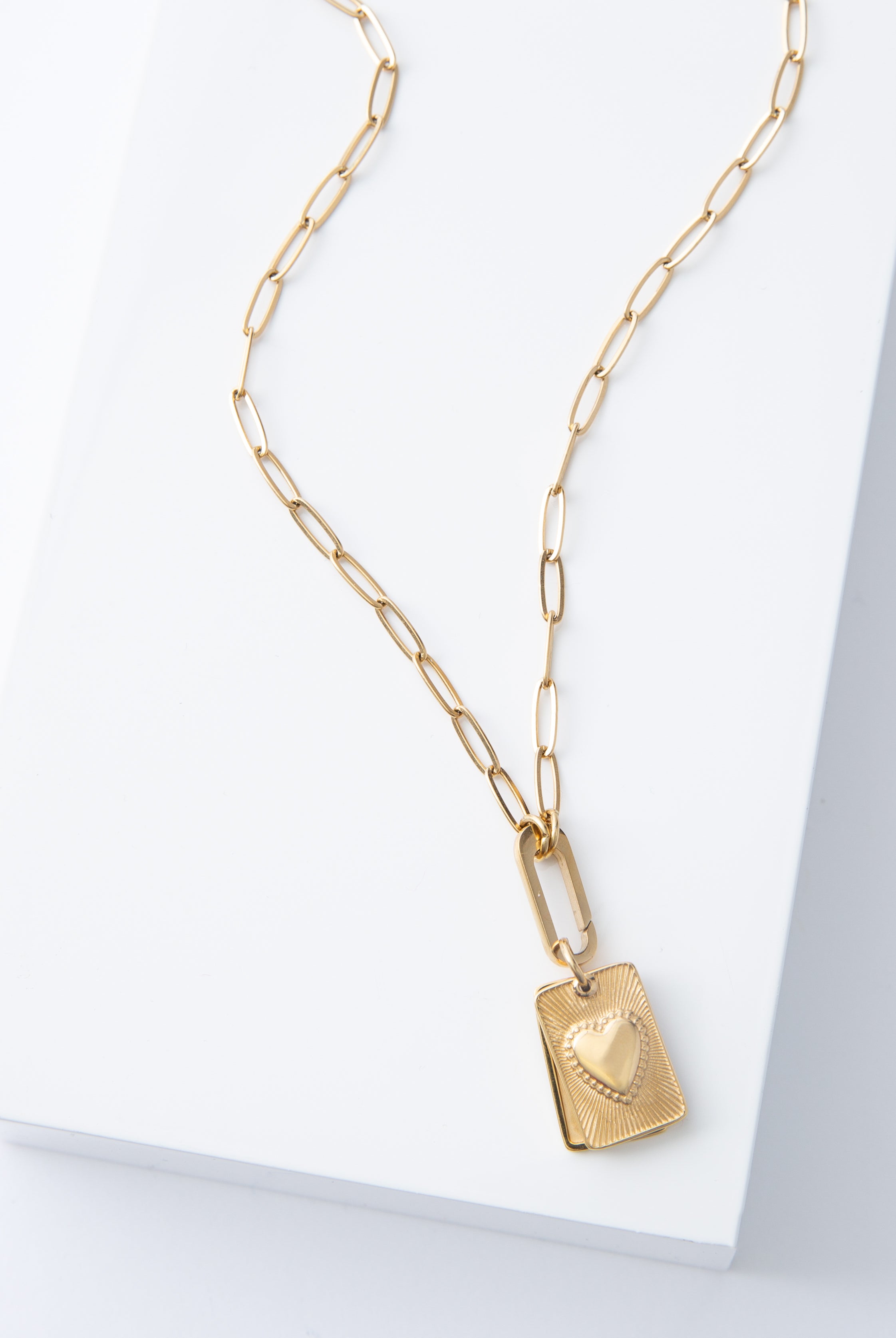 The Heart Keeper Necklace is a gold locket necklace with a gold chain composed of oval-shaped links. The locket pendant is suspended from a gold hoop. It is composed of two pieces of rectangular gold metal that can swing apart, allowing a photograph to be sandwiched between them. The piece on top has a raised heart design in the center.
