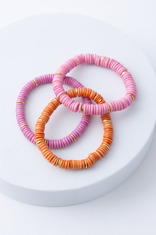The Heishi Bracelet Set includes three stretchy bracelets. The design of each bracelet is identical, but the colors vary. One is light lavender, one is light pink, and one is orange. The bracelets are composed of discs of shell strung together and accented with brass discs.