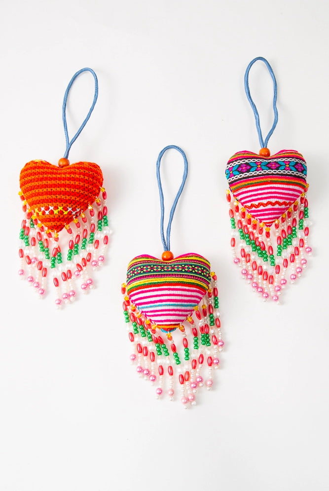 Three Hmong Heart Bag Charms are shown, demonstrating the textile variations that can occur due to the upcycled nature of the materials. While each generally has pink and red hues, the patterns and stripe colors vary on each charm.