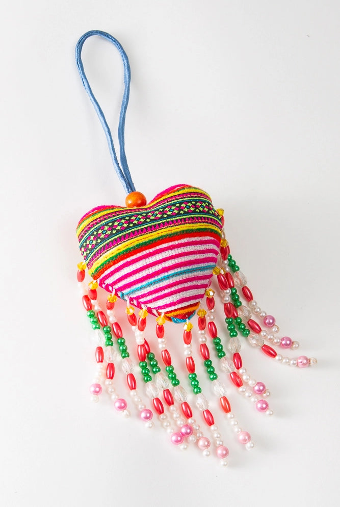 The Hmong Heart Bag Charm is a puffy heart charm made from traditional Vietnamese fabric. The brightly colored fabric is striped with rows of pink, white, blue, and patterned stripes. Beaded fringe hangs down below the heart. The charm has a fabric loop that allows it to be attached to a bag or other item.