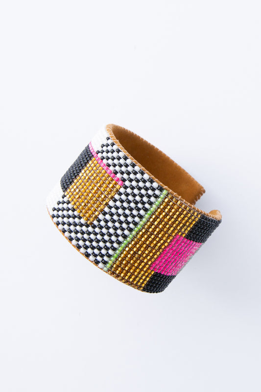 The Hologram Cuff is a wide leather cuff covered entirely in many rows of dainty glass beads. The beads are shades of black, white, hot pink, mint, and gold. They form a colorblocked, 80s-inspired pattern. The flexible cuff is open in the back, allowing it to slide onto the wrist.