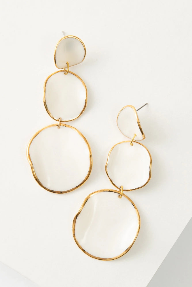 The Le Fantasia Earrings are chandelier drop earrings composed of a stack of three white porcelain discs outlined in shining gold. The disc attached to the post is the smallest, and each disc below is a little larger. The discs are not flat and have curves that give them a more organic look.