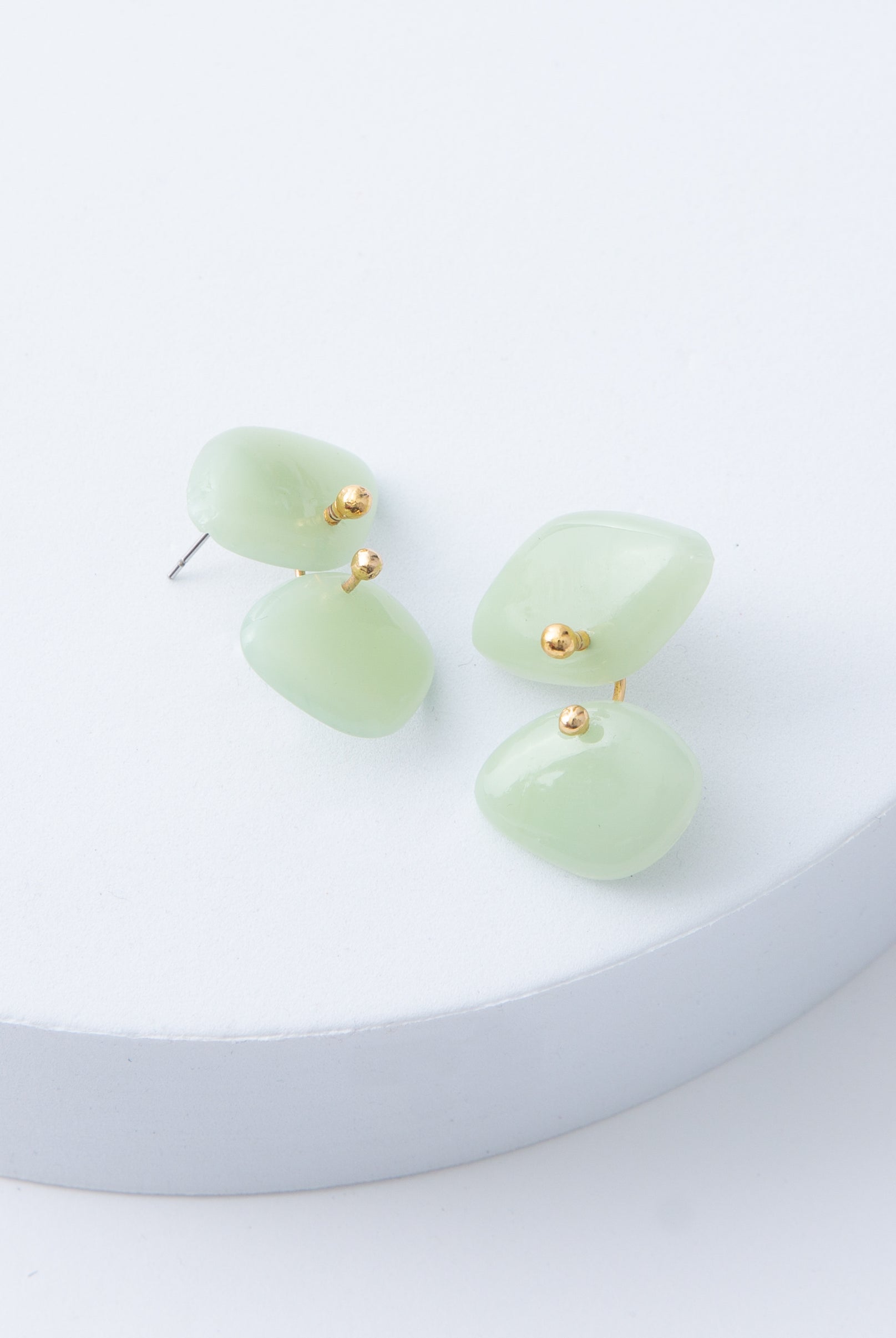 The Lilypad Earrings are post-style earrings with a minimal chandelier look. Two diamond-shaped glass beads in a light jade color are stacked on top of each other. The diamonds are connected by a single brass ring.