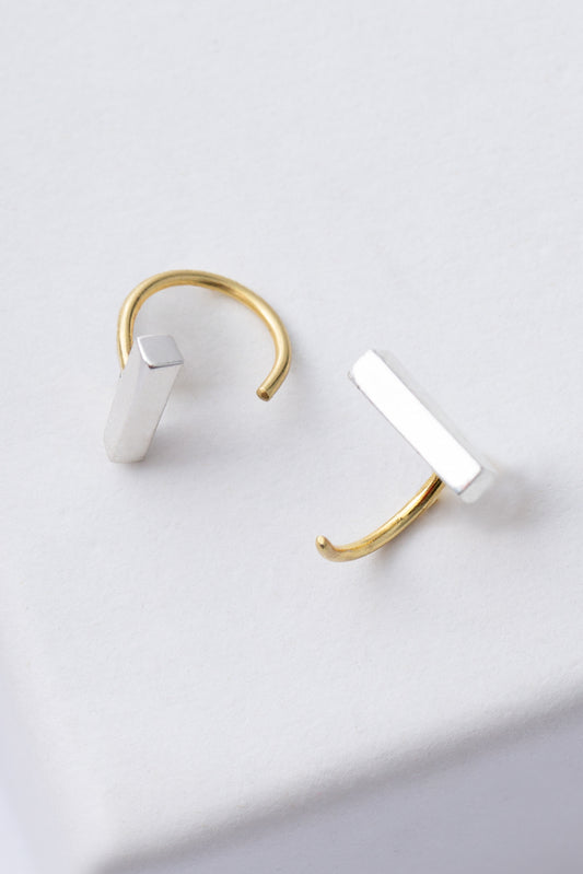 The Little Loop Earrings are gold, semi-hoop earrings that are open at the front. A silver bar sits at one end of the open hoop. The gold hoop slides through the piercing, and the silver bar rests against the front of the earlobe.