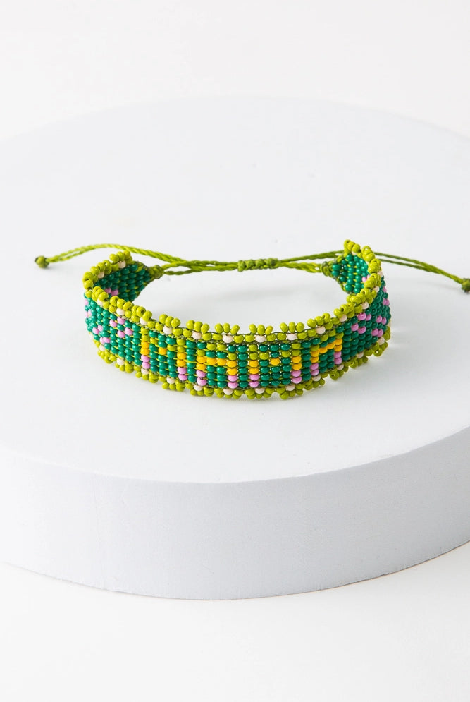 The Mama Beaded Bracelet is an adjustable thread bracelet featuring a band of green glass beads. Within the band, the word "Mama" is formed with lime, yellow, and pink glass beads. A row of lime glass beads forms a border around the bracelet. A lime thread closure at the back of the bracelet can be adjusted by pulling on both ends of the thread.