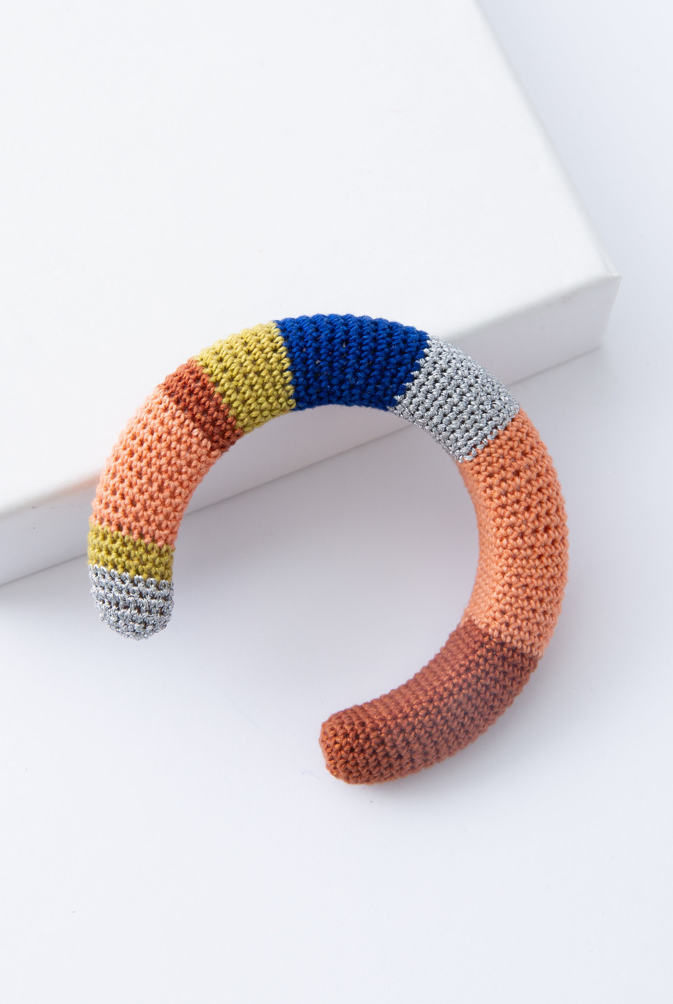 The Maza Crocheted Cuff is a rounded cuff covered entirely in crocheted thread. The thread color alternates between stripes of silver, mustard, peach, rust, and blue. The alternating stripes vary in width and create a color-blocked look. The cuff is open at the back to allow it to slide onto the wrist.