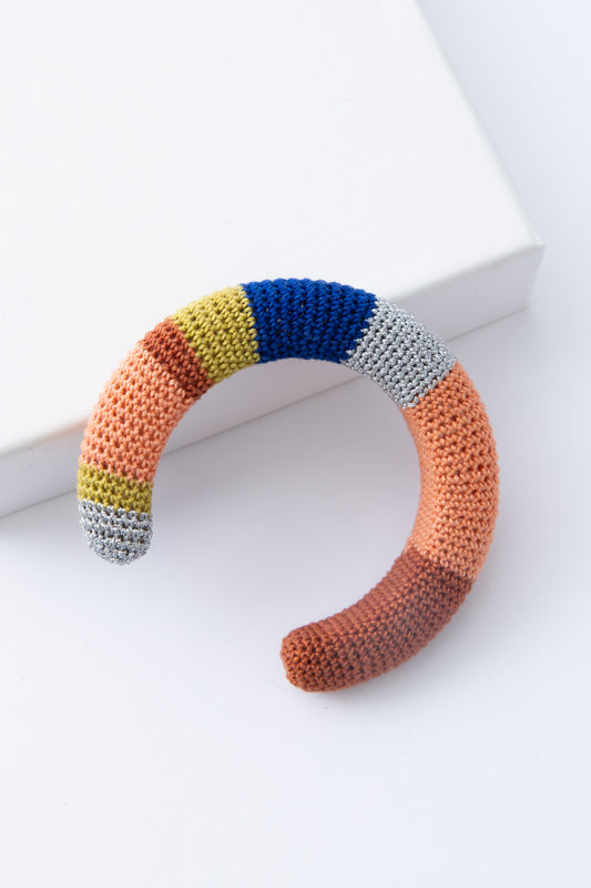 The Maza Crocheted Cuff is a rounded cuff covered entirely in crocheted thread. The thread color alternates between stripes of silver, mustard, peach, rust, and blue. The alternating stripes vary in width and create a color-blocked look. The cuff is open at the back to allow it to slide onto the wrist. 